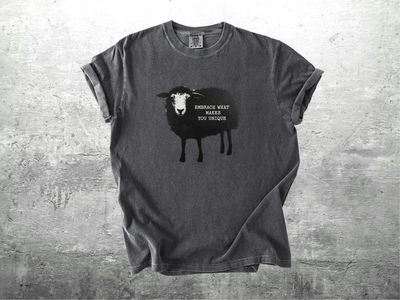 Puede incluir: Camiseta gris oscuro con una ilustraci&oacute;n de oveja negra y el texto "EMBRACE WHAT MAKES YOU UNIQUE." La camiseta tiene mangas enrolladas y cuello redondo. La camiseta est&aacute; hecha de un material suave y c&oacute;modo.