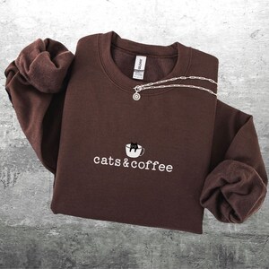 May include: A brown sweatshirt with the embroidered text "cats & coffee" and a graphic of a cat in a coffee cup. A silver necklace with a round pendant is draped across the top. The sweatshirt is folded on a gray surface.