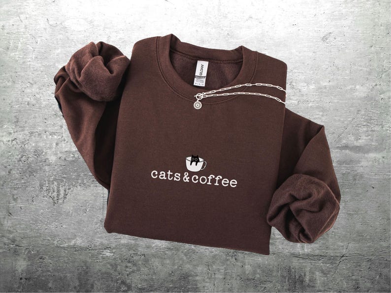 May include: A brown sweatshirt with the embroidered text "cats & coffee" and a graphic of a cat in a coffee cup. A silver necklace with a round pendant is draped across the top. The sweatshirt is folded on a gray surface.