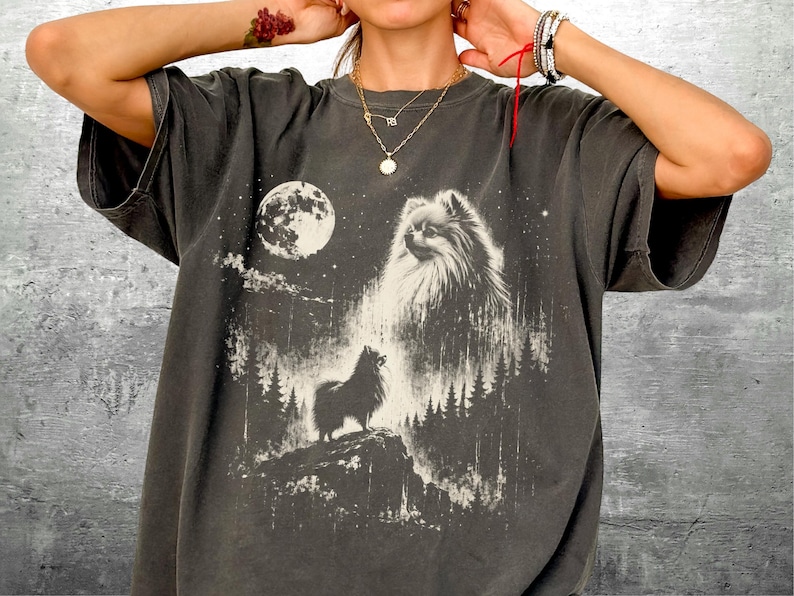 May include: Dark gray t-shirt featuring a white graphic of a Pomeranian dog under a full moon. The design includes a silhouette of a dog on a hill and a forest. The shirt has short sleeves and a relaxed fit.