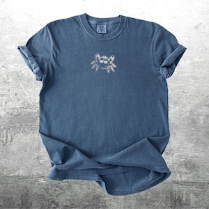 May include: A blue t-shirt with a small crab design on the front. The shirt has rolled-up sleeves and a crew neck. The crab design is a light color, contrasting with the darker blue of the shirt.
