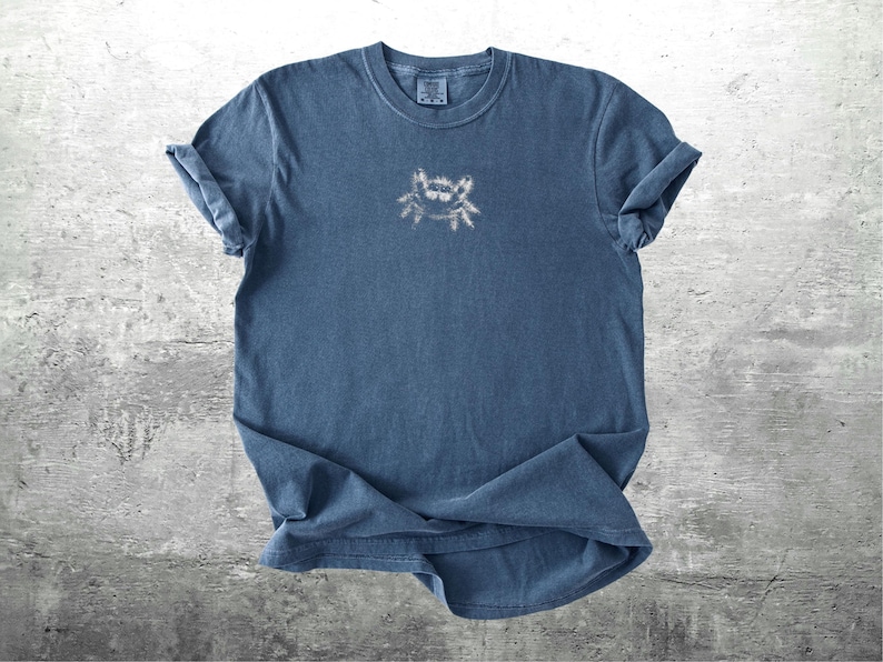 May include: A blue t-shirt with a small crab design on the front. The shirt has rolled-up sleeves and a crew neck. The crab design is a light color, contrasting with the darker blue of the shirt.