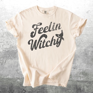 K&ouml;nnte beinhalten: Cremefarbenes T-Shirt mit dem Schriftzug "Feelin Witchy" in verblichener schwarzer Schrift. Ein Hexenhut-Grafik ist in das Design integriert. Das Shirt hat kurze &Auml;rmel und einen Rundhalsausschnitt.