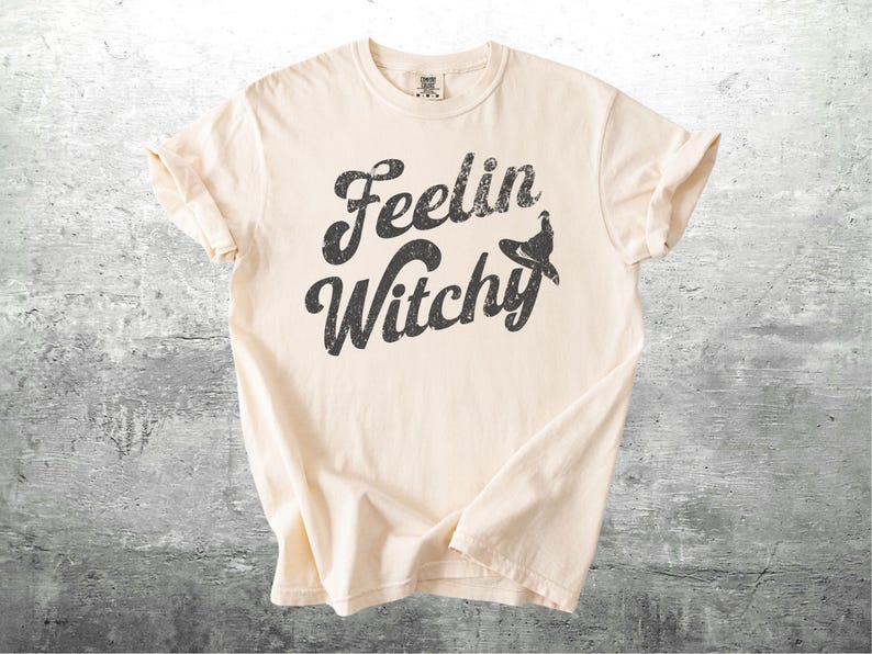 K&ouml;nnte beinhalten: Cremefarbenes T-Shirt mit dem Schriftzug "Feelin Witchy" in verblichener schwarzer Schrift. Ein Hexenhut-Grafik ist in das Design integriert. Das Shirt hat kurze &Auml;rmel und einen Rundhalsausschnitt.