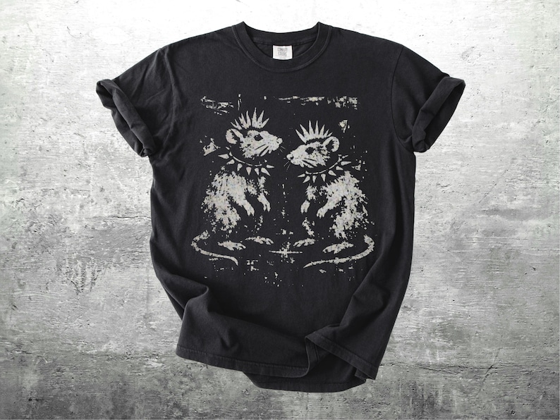 K&ouml;nnte beinhalten: Schwarzes T-Shirt mit einem verblichenen Aufdruck von zwei Ratten, die jeweils einen Stachelhalsband und eine Krone tragen. Die Ratten blicken sich an. Das Design hat eine verwaschene, Vintage-&Auml;sthetik. Die &Auml;rmel sind hochgekrempelt.