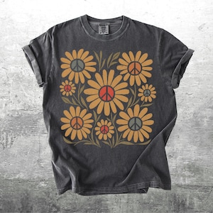 May include: A charcoal gray short-sleeve t-shirt featuring a graphic print of multiple yellow-orange daisies with green stems and leaves. Each flower center contains a peace sign symbol in shades of red, orange, or gray. The shirt has a crew neckline.
