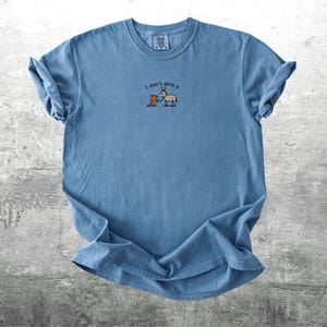 May include: Blue t-shirt with rolled sleeves. The shirt features the phrase "I don't give a" above a cartoon illustration of a bear and a donkey. The shirt is made of a soft material and is laid flat on a grey surface.