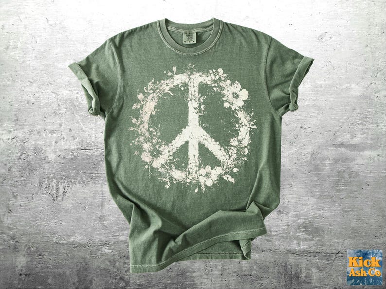 May include: A sage green short-sleeve t-shirt featuring a large, distressed white graphic of a peace sign intertwined with floral and leaf patterns centered on the chest. The shirt has a crew neckline and a relaxed, casual fit with rolled sleeves.