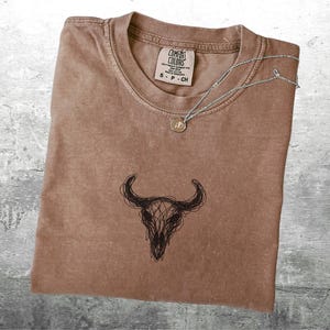 May include: A brown Comfort Colors t-shirt with a black bull skull graphic. A silver necklace with a small pendant rests on the shirt. The shirt has a small tag that reads "S - P - CH".