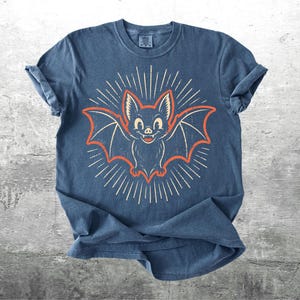 May include: A blue short-sleeve t-shirt featuring a central graphic of a smiling bat with orange outlines and white radiating sunburst lines. The bat has large ears and visible fangs. The shirt has a crew neckline and a slightly faded, textured appearance.