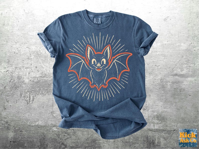 May include: A blue short-sleeve t-shirt featuring a central graphic of a smiling bat with orange outlines and white radiating sunburst lines. The bat has large ears and visible fangs. The shirt has a crew neckline and a slightly faded, textured appearance.