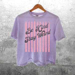 May include: A light purple short-sleeve t-shirt featuring a graphic design of vertical pink stripes and the dark grey text "Be Kind Stay Weird" printed in a cursive font across the centre. The shirt is displayed on a wooden hanger.