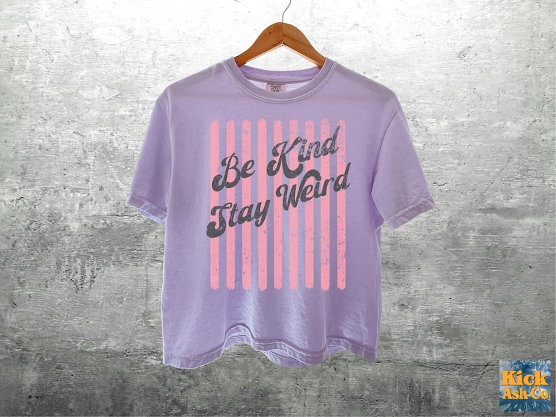 May include: A light purple short-sleeve t-shirt featuring a graphic design of vertical pink stripes and the dark grey text "Be Kind Stay Weird" printed in a cursive font across the centre. The shirt is displayed on a wooden hanger.