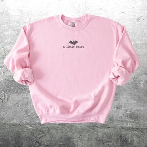 May include: A light pink sweatshirt with rolled-up sleeves. The text "a little batty" is printed in black, along with a bat graphic. The sweatshirt is made of a soft material and is suitable for casual wear.
