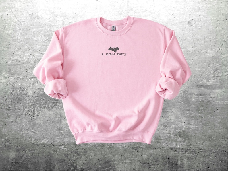 May include: A light pink sweatshirt with rolled-up sleeves. The text "a little batty" is printed in black, along with a bat graphic. The sweatshirt is made of a soft material and is suitable for casual wear.