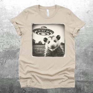 May include: A beige t-shirt with a black and white image of a possum looking up at a flying saucer. The image is in a vintage style.