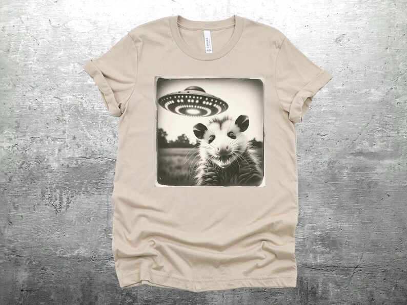 May include: A beige t-shirt with a black and white image of a possum looking up at a flying saucer. The image is in a vintage style.