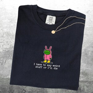 May include: Dark navy t-shirt featuring a cartoon frog in a pink bunny suit and yellow boots. The text reads "I have to say weird stuff or I'll die". A gold necklace with a pendant is draped on the shirt.
