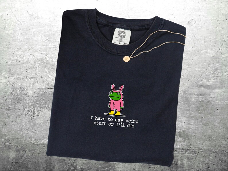 May include: Dark navy t-shirt featuring a cartoon frog in a pink bunny suit and yellow boots. The text reads "I have to say weird stuff or I'll die". A gold necklace with a pendant is draped on the shirt.