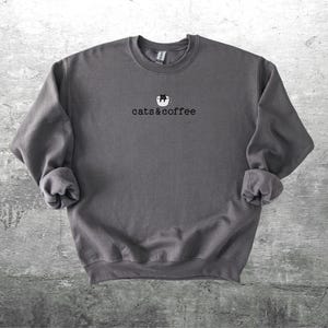 May include: A dark gray sweatshirt with the text "cats & coffee" in a simple font. Above the text is a small graphic of a cat in a coffee cup. The sweatshirt has a crew neck and long sleeves with rolled cuffs.