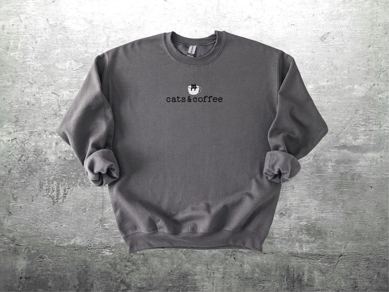 May include: A dark gray sweatshirt with the text "cats & coffee" in a simple font. Above the text is a small graphic of a cat in a coffee cup. The sweatshirt has a crew neck and long sleeves with rolled cuffs.