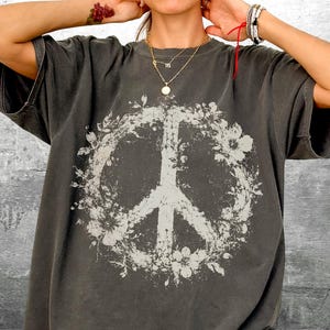 May include: A charcoal gray short-sleeve t-shirt featuring a large, distressed white graphic of a peace sign encircled by floral elements. The design has a weathered, vintage-inspired texture printed on the front center of the garment.