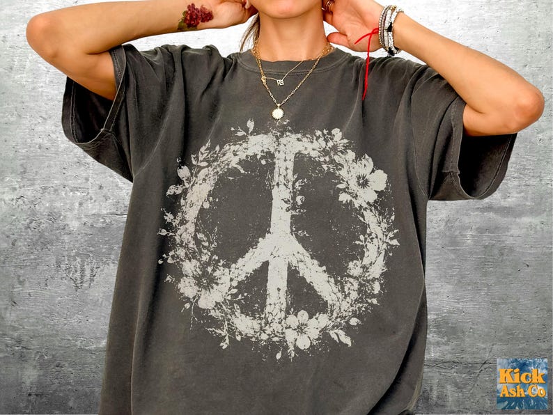May include: A charcoal gray short-sleeve t-shirt featuring a large, distressed white graphic of a peace sign encircled by floral elements. The design has a weathered, vintage-inspired texture printed on the front center of the garment.