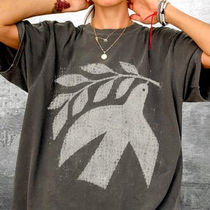 May include: A charcoal gray short-sleeve t-shirt featuring a distressed, light gray graphic print of a bird carrying an olive branch. The shirt has a crew neckline and a relaxed fit, with the text Kick Ash Co. visible in the bottom right corner.