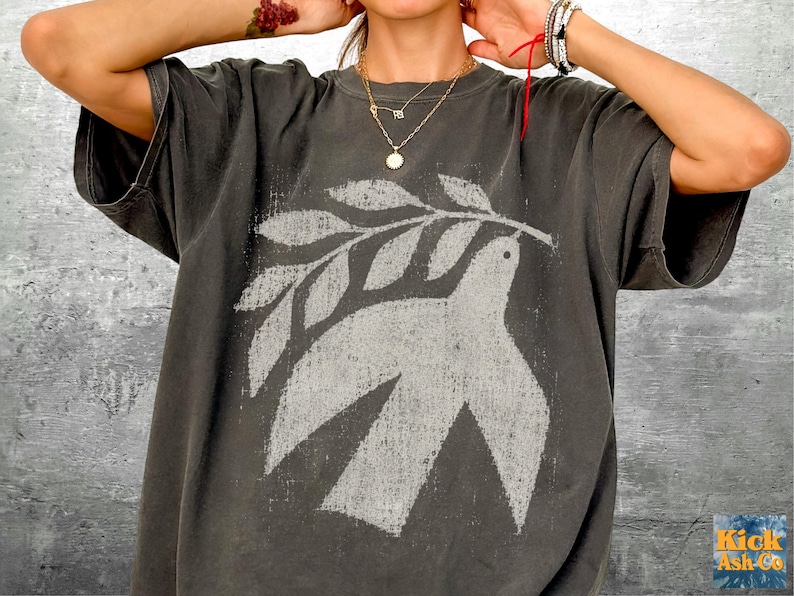 May include: A charcoal gray short-sleeve t-shirt featuring a distressed, light gray graphic print of a bird carrying an olive branch. The shirt has a crew neckline and a relaxed fit, with the text Kick Ash Co. visible in the bottom right corner.