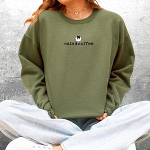 May include: An olive green sweatshirt with the text "cats & coffee" in a black, simple font. Above the text is a small graphic of a cat in a coffee cup. The sweatshirt is a solid color and has a crew neck.