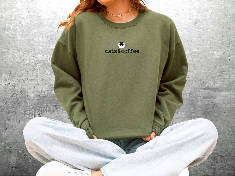 May include: An olive green sweatshirt with the text "cats & coffee" in a black, simple font. Above the text is a small graphic of a cat in a coffee cup. The sweatshirt is a solid color and has a crew neck.