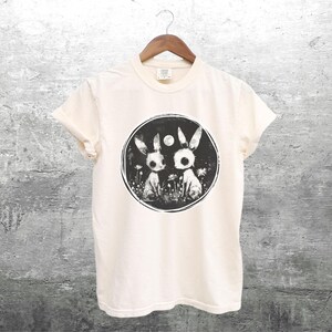 Op de afbeelding: Een wit T-shirt met een zwart-witte grafische print van twee konijnen in een cirkel. De konijnen kijken elkaar aan en hebben grote, ronde ogen. De cirkel is omringd door een ring van kleine bloemen.