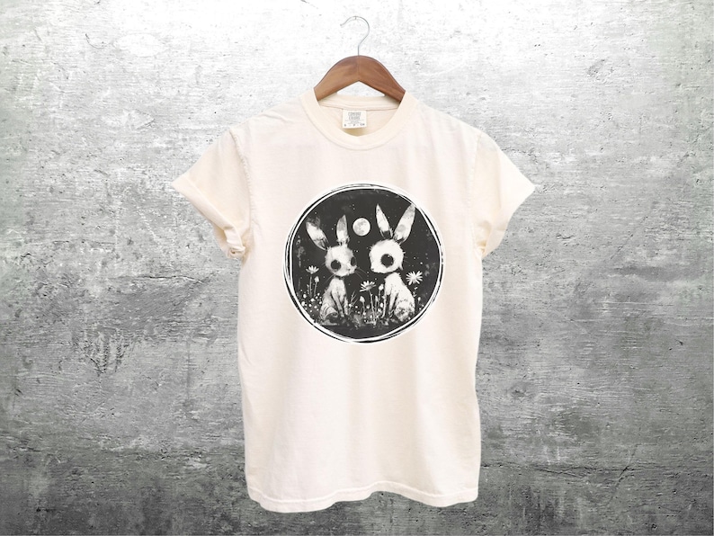 Op de afbeelding: Een wit T-shirt met een zwart-witte grafische print van twee konijnen in een cirkel. De konijnen kijken elkaar aan en hebben grote, ronde ogen. De cirkel is omringd door een ring van kleine bloemen.