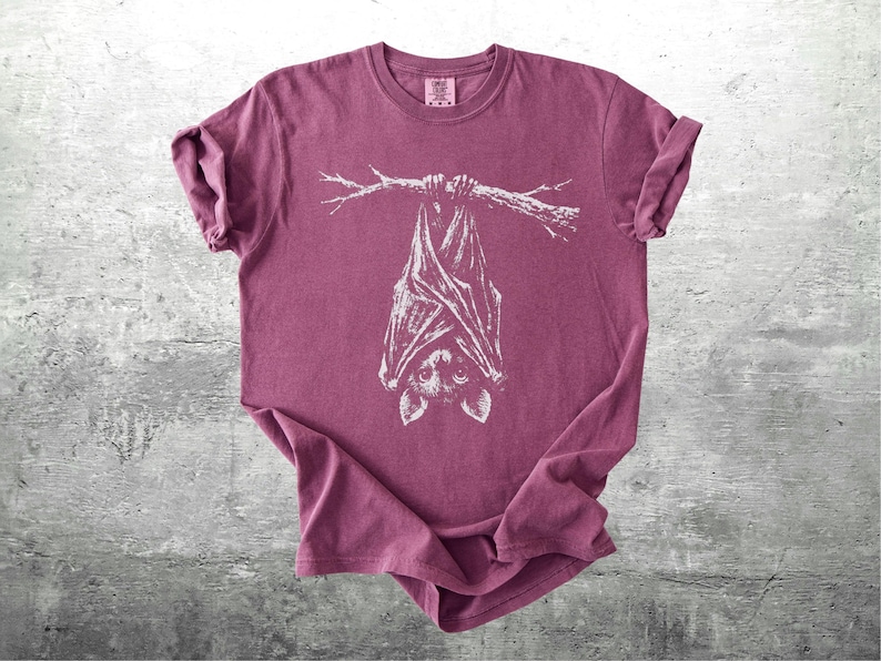 Op de afbeelding: Een bordeauxrood t-shirt met een witte illustratie van een vleermuis die aan een tak hangt. De vleugels en het gezicht van de vleermuis zijn duidelijk zichtbaar. Het shirt is opgevouwen, met opgerolde mouwen.