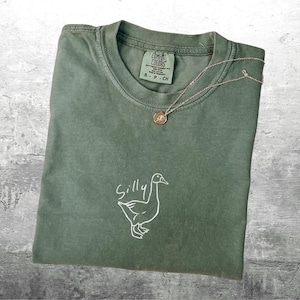May include: A green t-shirt with a white outline of a goose and the word "Silly" written above it.