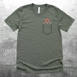 May include: A green t-shirt with a pocket featuring a raccoon wearing a red hat. The raccoon is peeking out of the pocket.