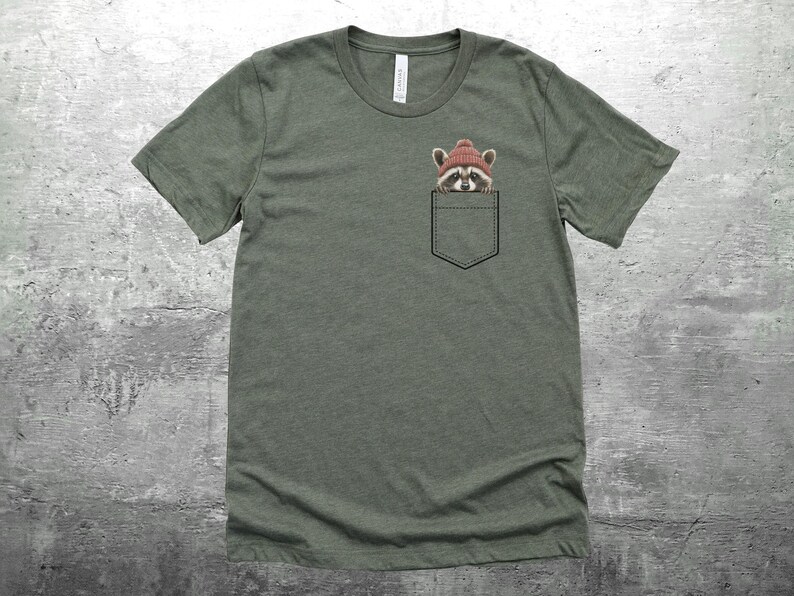 May include: A green t-shirt with a pocket featuring a raccoon wearing a red hat. The raccoon is peeking out of the pocket.