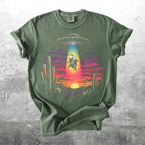 May include: An olive green crew neck t-shirt featuring a graphic print of a possum being abducted by a UFO in a desert landscape at sunset. The design includes cacti, a bright yellow sun, and a colorful beam of light against a purple sky.