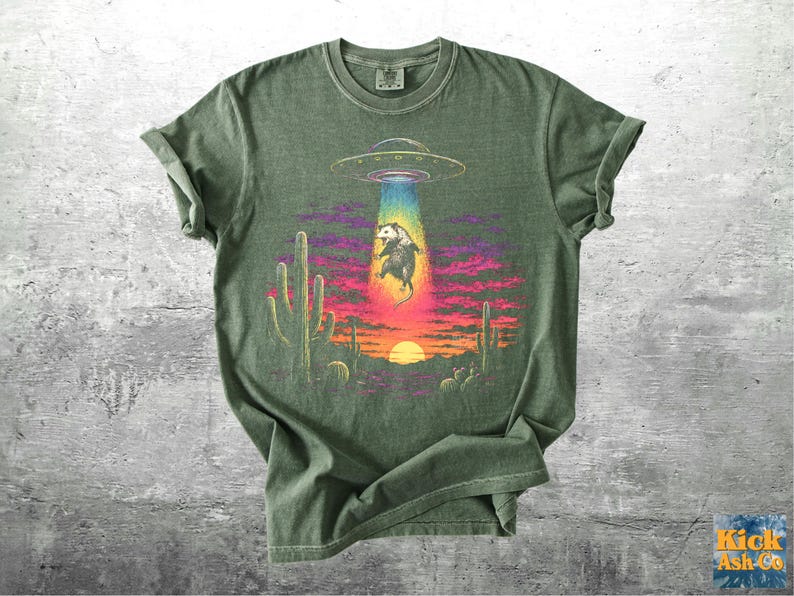 May include: An olive green crew neck t-shirt featuring a graphic print of a possum being abducted by a UFO in a desert landscape at sunset. The design includes cacti, a bright yellow sun, and a colorful beam of light against a purple sky.