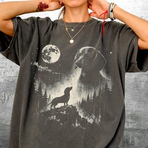 May include: Dark gray t-shirt featuring a dachshund dog design under a full moon. The graphic includes a silhouette of the dog on a hill, a larger dog portrait, and a forest scene. The shirt has short sleeves and a relaxed fit.