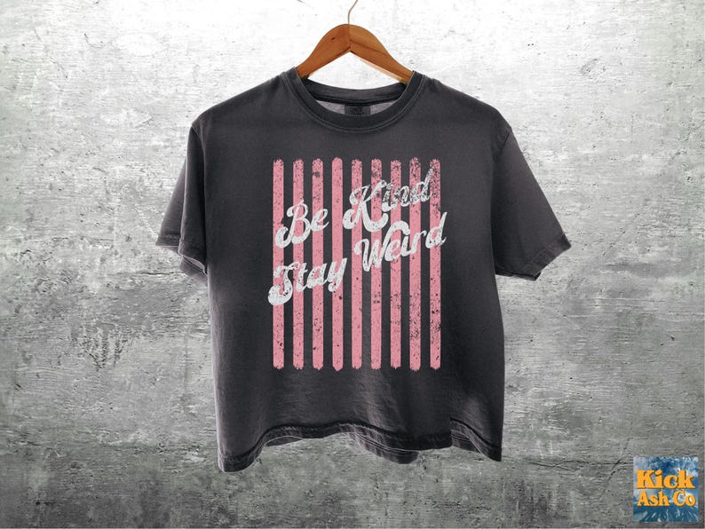 May include: A charcoal grey short-sleeve t-shirt featuring a graphic design of vertical pink stripes with a distressed texture. The white text printed over the stripes reads "Be Kind Stay Weird" in a cursive font. The shirt hangs on a wooden hanger.