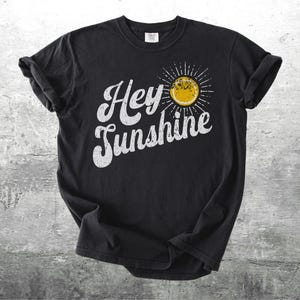 May include: Black t-shirt with the words "Hey Sunshine" in white, stylized lettering. A yellow sun graphic with radiating lines is on the right side of the text. The shirt is displayed on a textured gray surface.