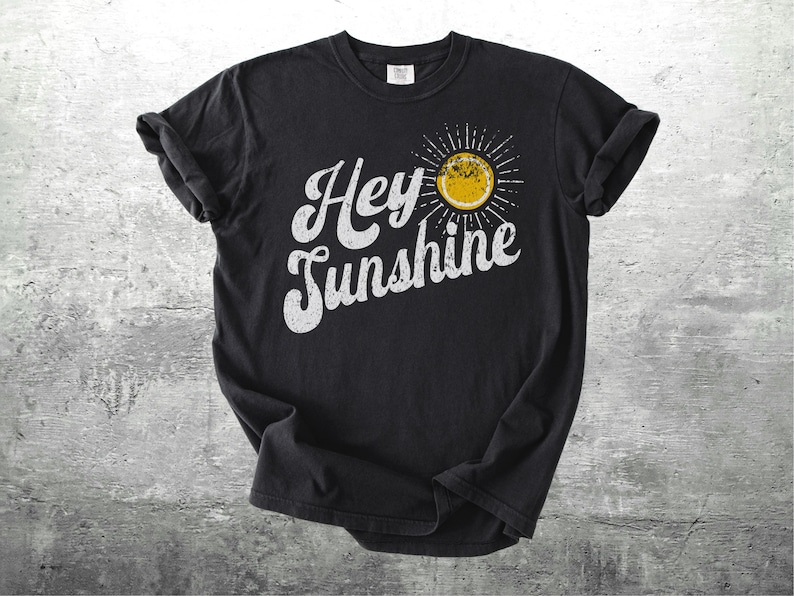 May include: Black t-shirt with the words "Hey Sunshine" in white, stylized lettering. A yellow sun graphic with radiating lines is on the right side of the text. The shirt is displayed on a textured gray surface.