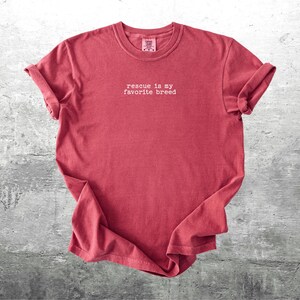 May include: A red t-shirt with rolled-up sleeves. The shirt has the phrase "rescue is my favorite breed" printed in white, centered on the front. The shirt is displayed on a gray, textured surface.