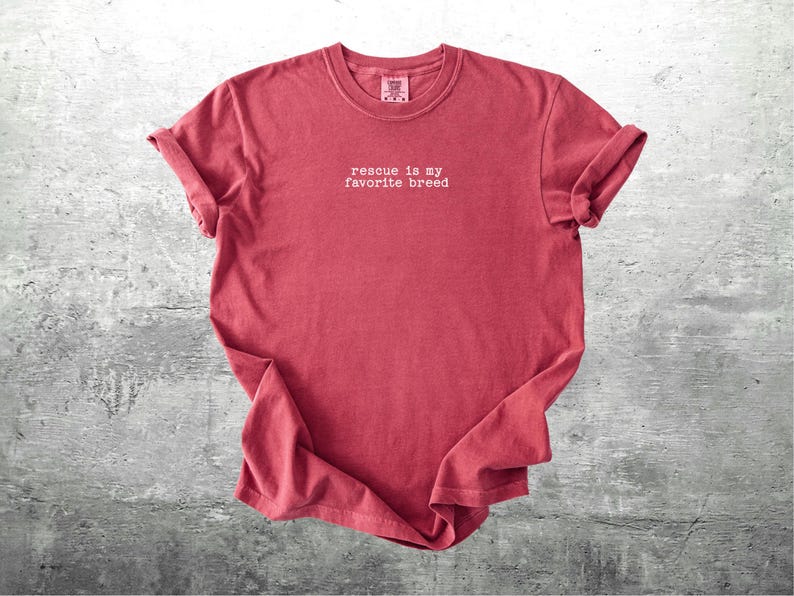 May include: A red t-shirt with rolled-up sleeves. The shirt has the phrase "rescue is my favorite breed" printed in white, centered on the front. The shirt is displayed on a gray, textured surface.