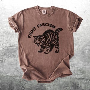 K&ouml;nnte beinhalten: Braunes T-Shirt mit dem Text "FIGHT FASCISM" &uuml;ber einer schwarzen Illustration einer w&uuml;tenden Katze. Die Katze hat Streifen und einen ge&ouml;ffneten Mund. Das T-Shirt ist gefaltet und hat einen Vintage-Look.