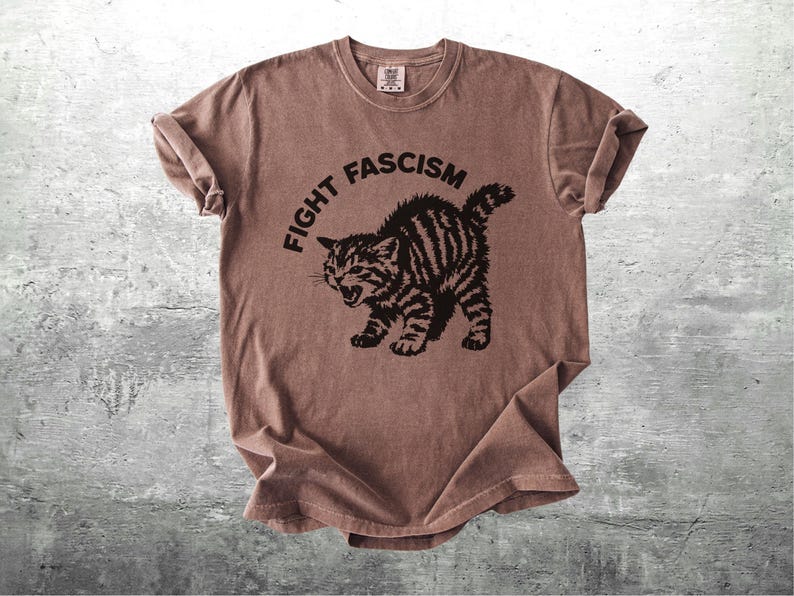 K&ouml;nnte beinhalten: Braunes T-Shirt mit dem Text "FIGHT FASCISM" &uuml;ber einer schwarzen Illustration einer w&uuml;tenden Katze. Die Katze hat Streifen und einen ge&ouml;ffneten Mund. Das T-Shirt ist gefaltet und hat einen Vintage-Look.
