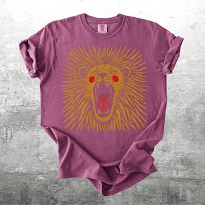 May include: A maroon t-shirt with a gold lion graphic, open-mouthed and red-eyed. The lion's mane has radiating lines. The shirt is displayed on a grey, textured surface.