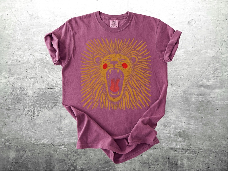 May include: A maroon t-shirt with a gold lion graphic, open-mouthed and red-eyed. The lion's mane has radiating lines. The shirt is displayed on a grey, textured surface.