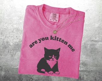 Puede incluir: Camiseta rosa con la frase "are you kitten me" y un dibujo de gato en blanco y negro. Un collar dorado con un colgante está colocado sobre la camiseta.